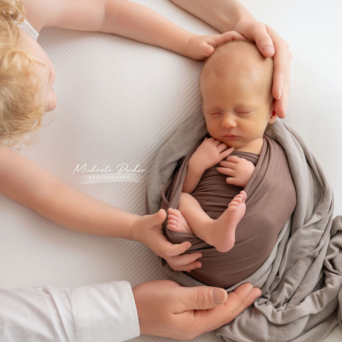 Schlafendes Neugeborenes, eingewickelt in braun-graue Tücher, wird liebevoll von mehreren Händen – den Händen der Eltern und des Geschwisterkindes – gehalten und umrahmt.